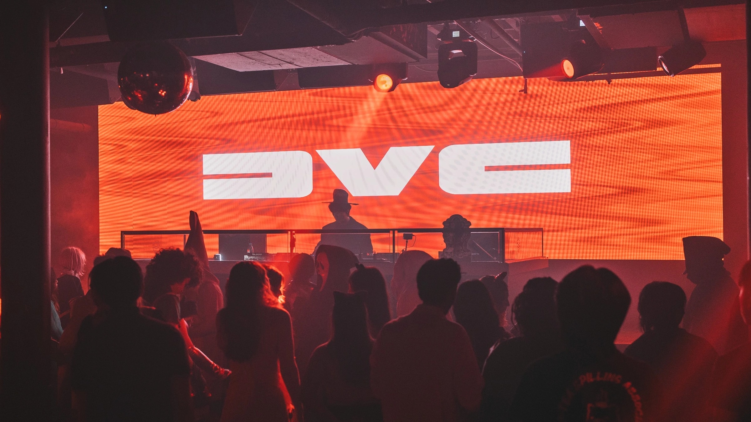 A crowd facing a stage at Bloom Nightclub, with the DVC logo glowing across a wall of orange LED light.